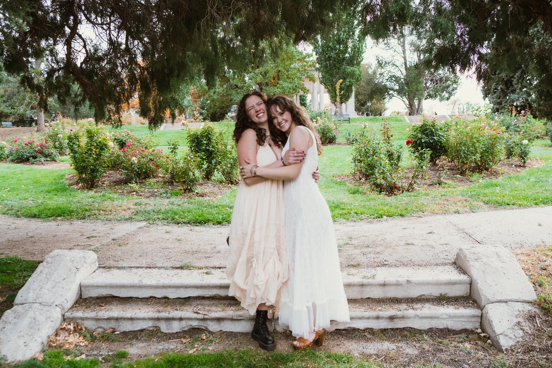 Two friends hugging and laughing together on stone steps in a rose garden